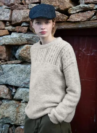 woman standing in front of stone house wearing an off-white eyelet stitch woolen sweater with a tweed cap